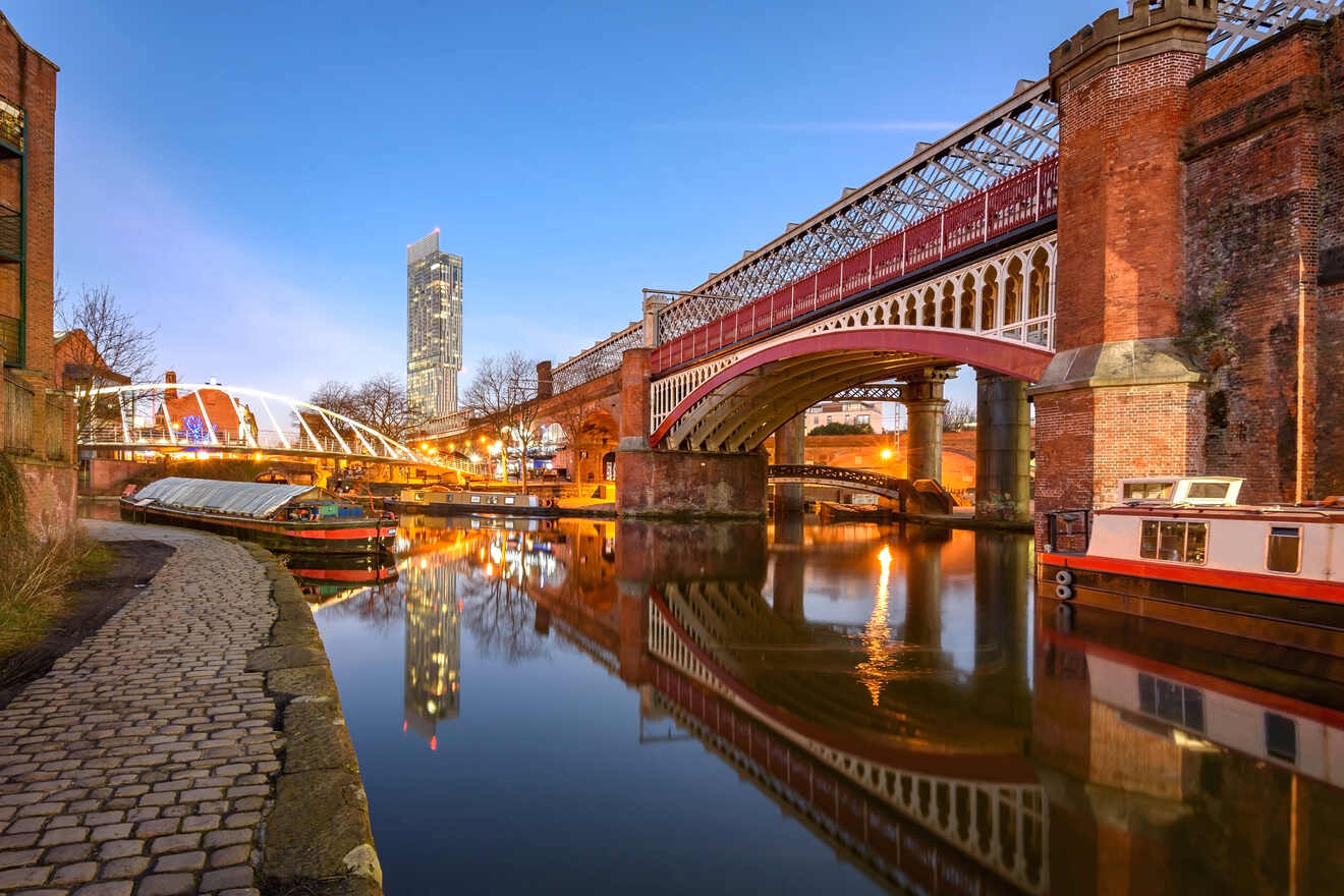 Dusk scene at Manchester's Castlefield district with historic bridges over calm canals, reflecting the lights and modern skyscrapers in the water